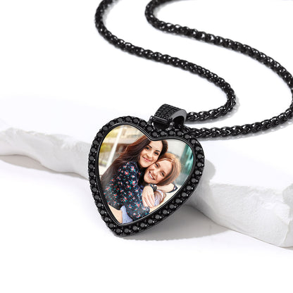 A heart-shaped pendant necklace with a black bezel set with small clear stones, featuring a custom photo insert of two women hugging. The necklace is displayed on a white textured stone prop against a clean white background.