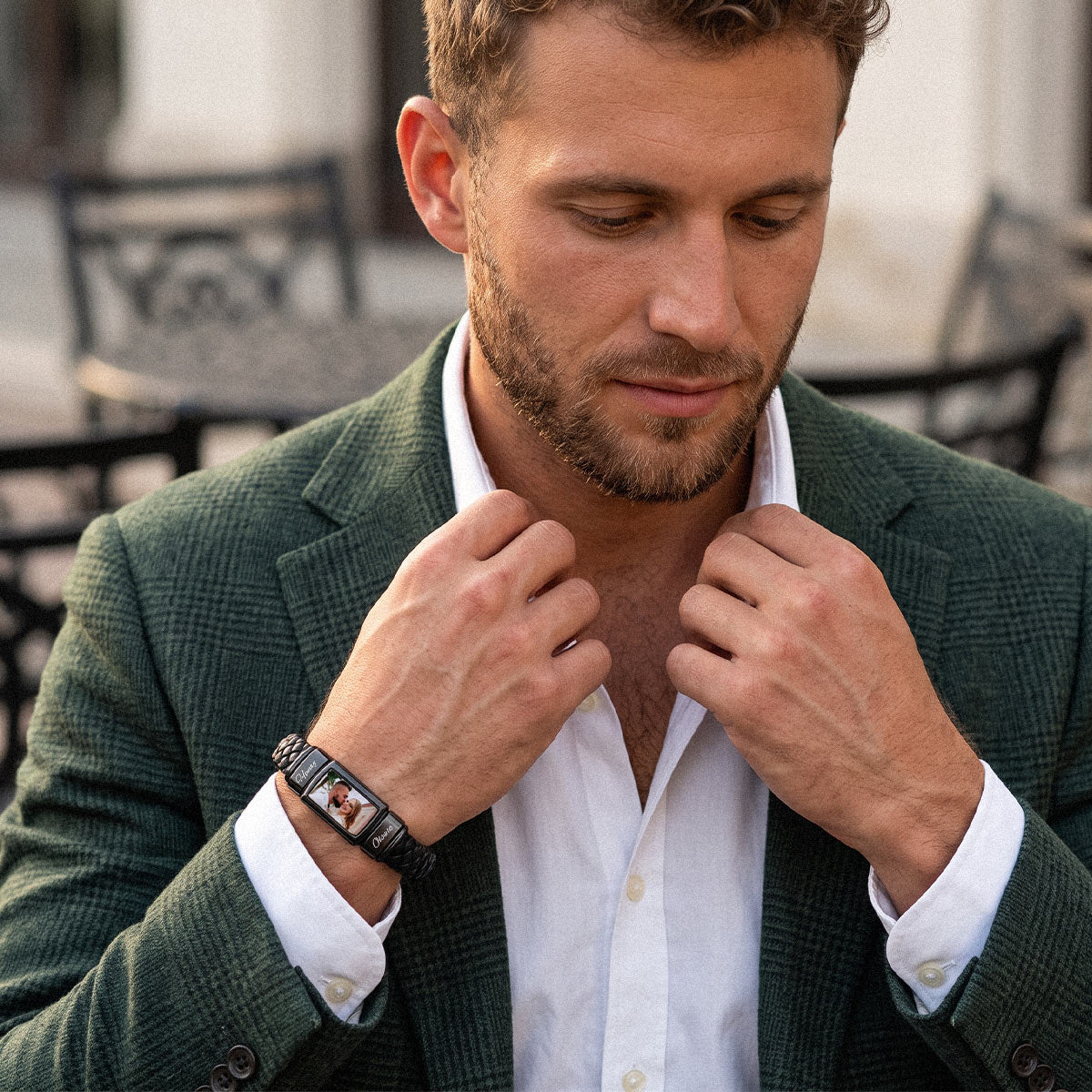 A man wearing a dark green textured blazer and white dress shirt, looking down while adjusting his collar. He is wearing a black custom photo leather bracelet with a rectangular digital watch face on his wrist.