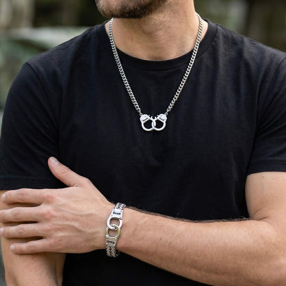 A man wearing a silver chain necklace with a double-ring pendant and a matching silver link bracelet, styled with a black t-shirt against a blurred outdoor background.