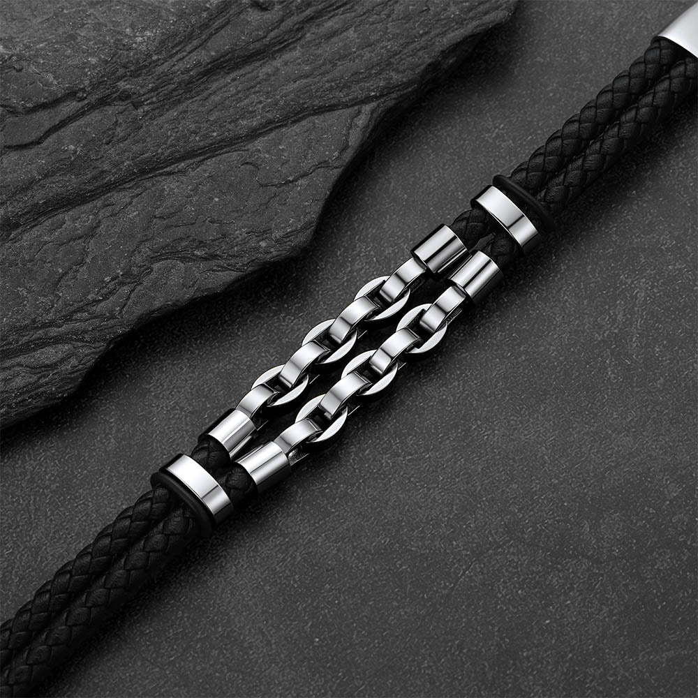 A men's bracelet featuring a black braided leather strap and a central silver-toned chain link design, displayed diagonally on a dark textured slate background.