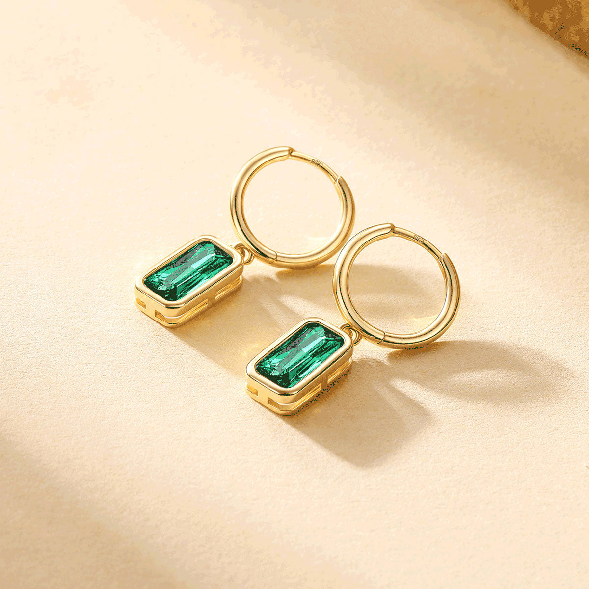A pair of gold May birthstone hoop earrings with rectangular green gemstone charms, displayed on a warm beige textured surface.