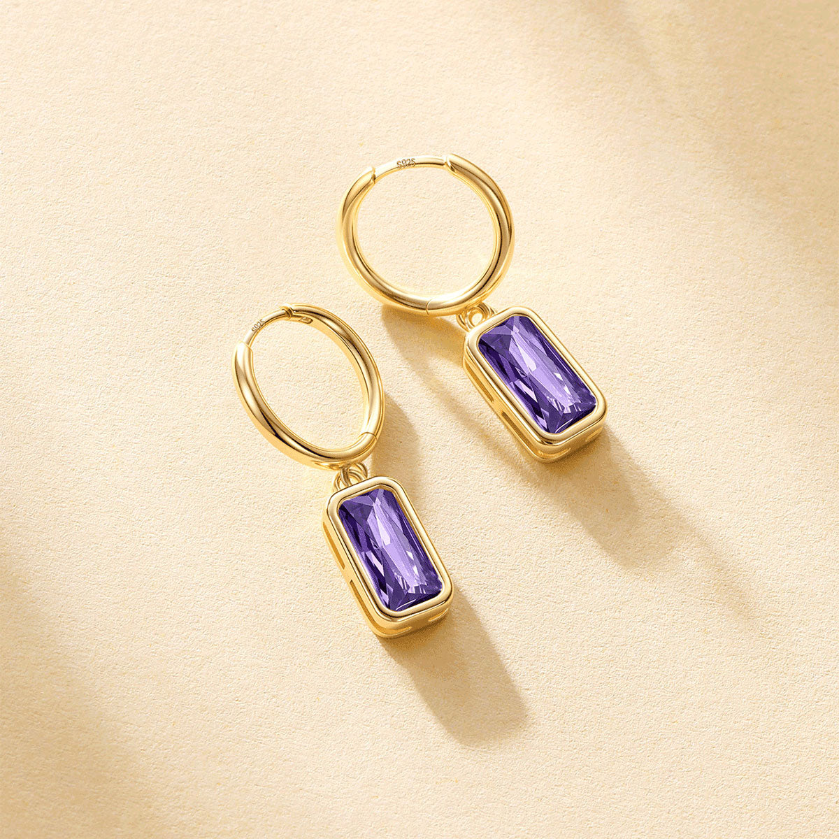 A pair of gold birthstone hoop earrings with rectangular purple gemstones, displayed on a textured beige surface with soft shadows.
