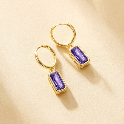 A pair of gold birthstone hoop earrings with rectangular purple gemstones, displayed on a textured beige surface with soft shadows.