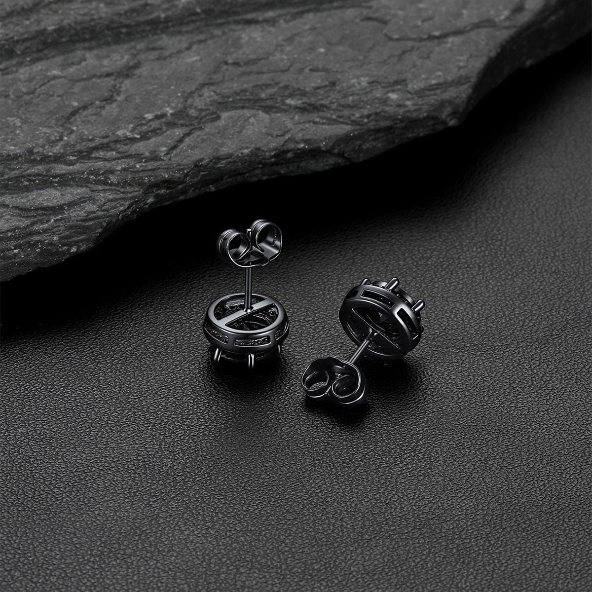A pair of matte black CZ stud earrings, displayed on a dark textured surface with a slate stone in the background.