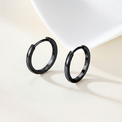 A pair of matte black huggie earrings with a 925 sterling silver hallmark engraved on the inside, displayed on a clean, light beige surface with a white curved prop in the background.