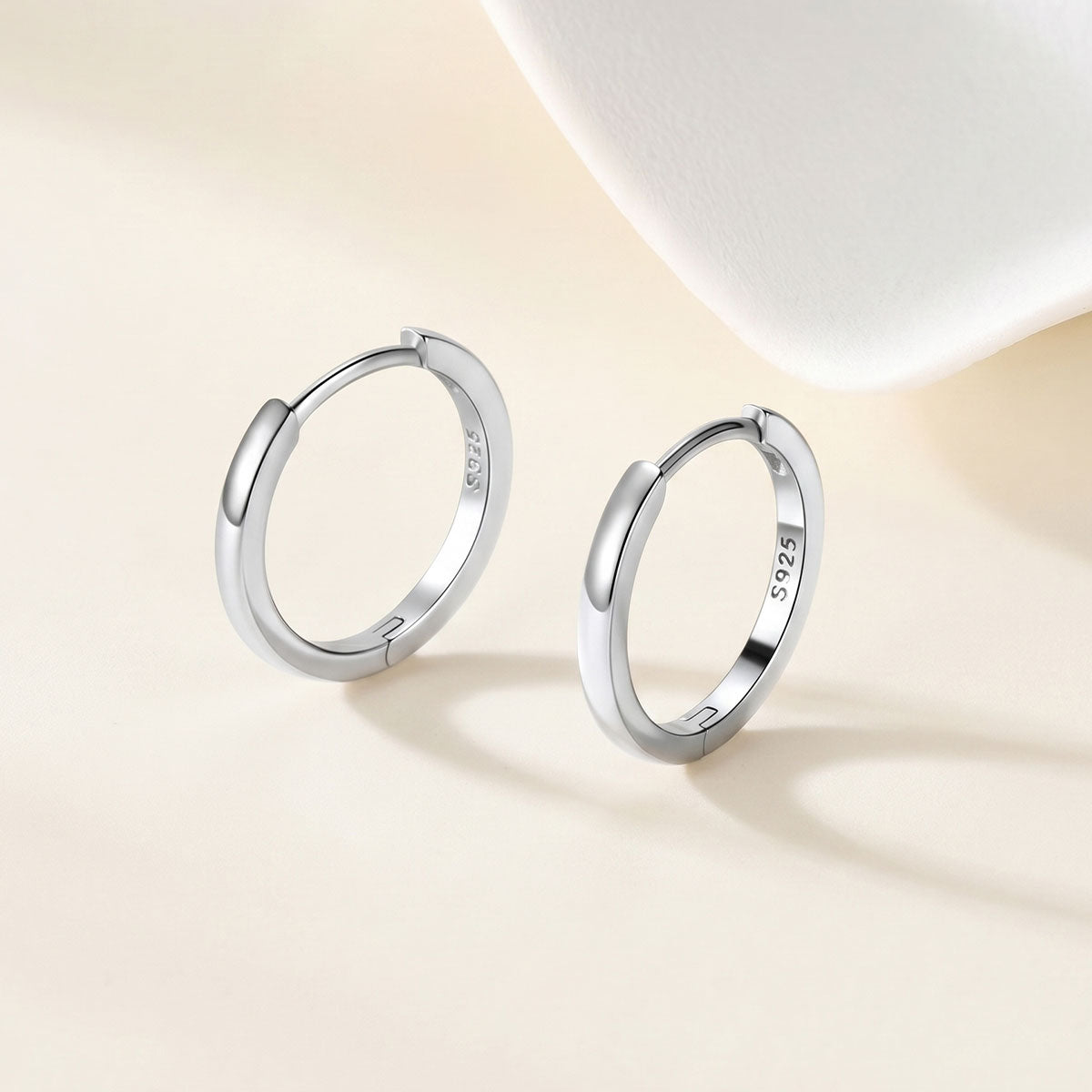 A pair of polished silver huggie earrings with a 925 sterling silver hallmark, displayed on a warm beige surface with a soft white decorative prop in the background.