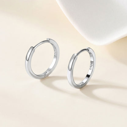 A pair of polished silver huggie earrings with a 925 sterling silver hallmark, displayed on a warm beige surface with a soft white decorative prop in the background.