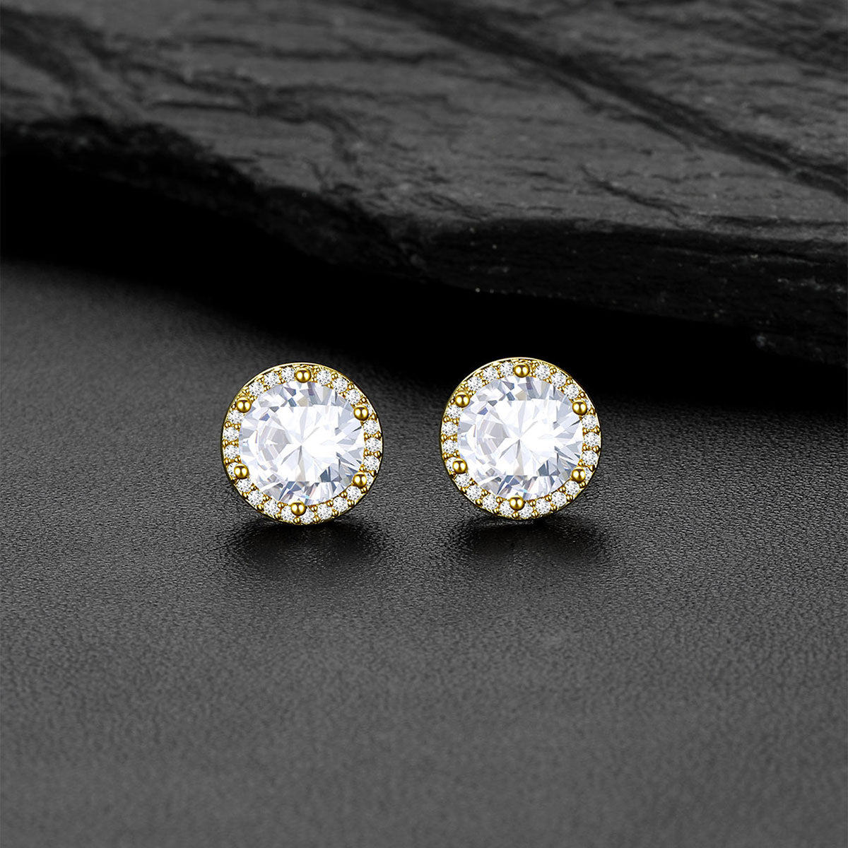 A pair of round CZ stud earrings with large clear center stones surrounded by a halo of small clear stones in yellow gold settings, displayed on a dark textured surface with a black slate prop.