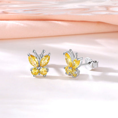 A pair of silver butterfly stud earrings with yellow marquise-cut and round-cut stones, displayed on a soft white surface with a peach-colored satin fabric in the background.