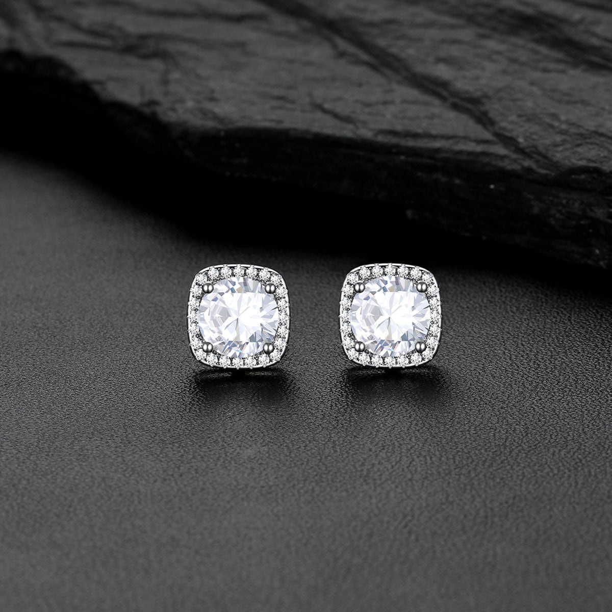 A pair of square cushion-cut CZ stud earrings with clear gemstones in a silver-toned setting, displayed on a dark textured surface with a black slate background.