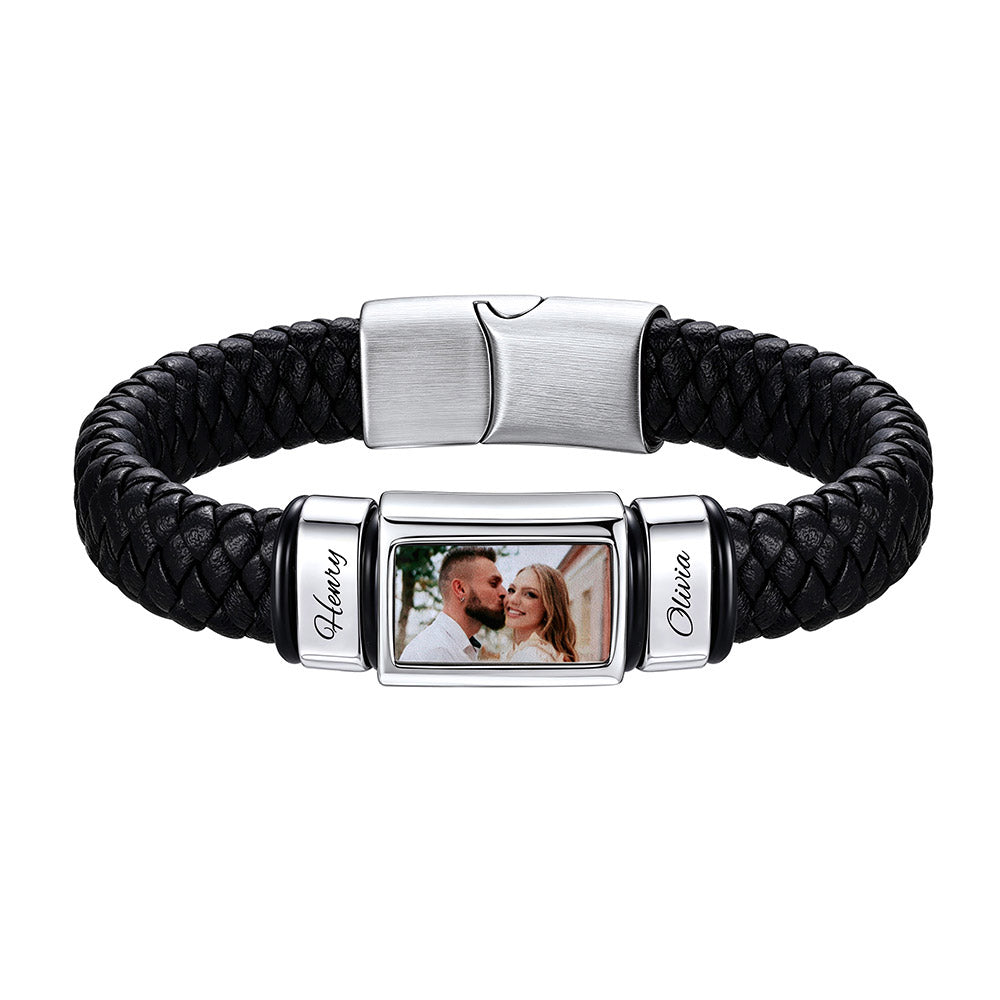 A personalized black braided leather bracelet with a silver-toned metal clasp. The center of the bracelet features a rectangular photo frame showing a couple, with the names 'Henry' and 'Olivia' engraved on the silver side plates.