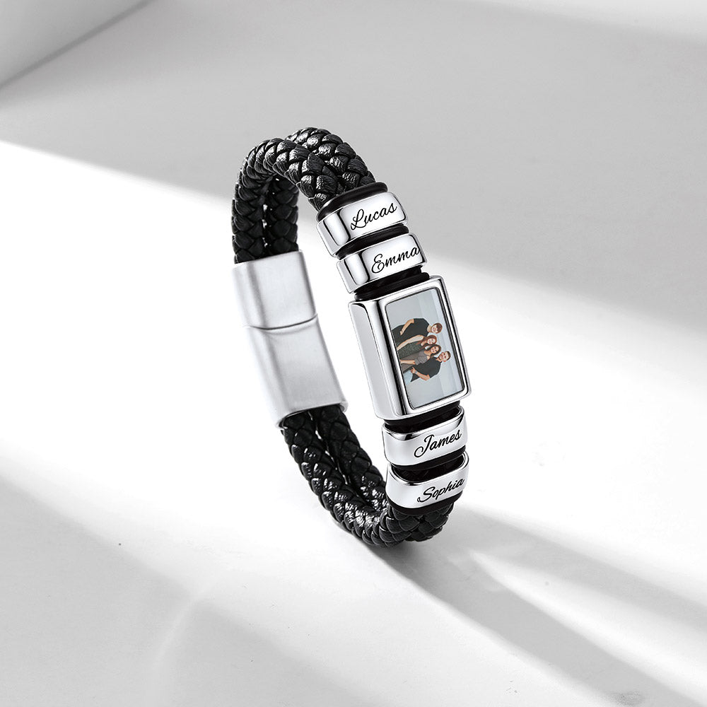 A personalized black braided leather bracelet with silver-tone metal charms. The bracelet features a rectangular charm engraved with a cartoon character and the name 'Emma', flanked by round silver charms engraved with 'Lions', 'James', and 'Saudade'.
