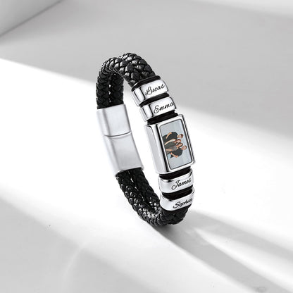 A personalized black braided leather bracelet with silver-tone metal charms. The bracelet features a rectangular charm engraved with a cartoon character and the name 'Emma', flanked by round silver charms engraved with 'Lions', 'James', and 'Saudade'.