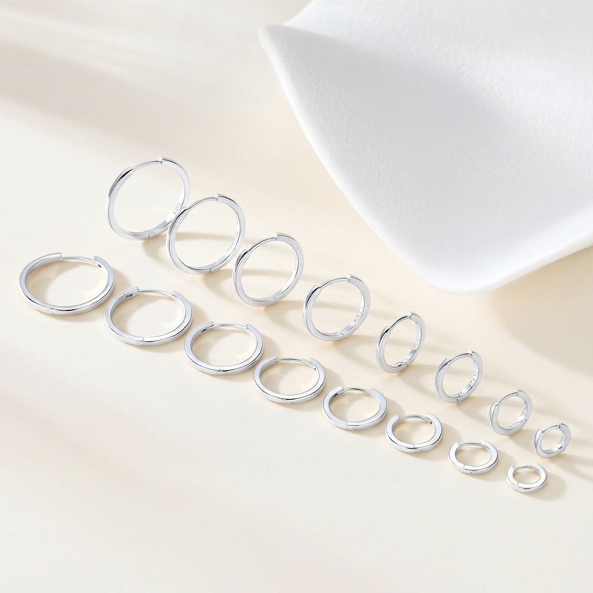 A set of silver huggie earrings in multiple sizes, arranged diagonally on a light beige surface with a white textured prop in the background.