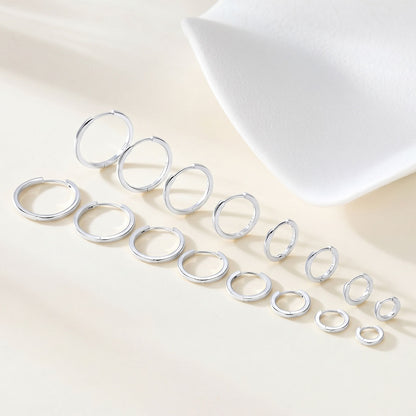 A set of silver huggie earrings in multiple sizes, arranged diagonally on a light beige surface with a white textured prop in the background.
