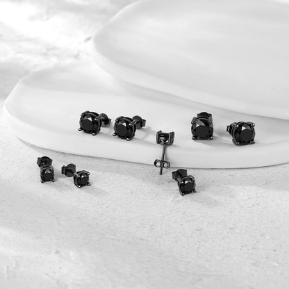 A set of small black CZ stud earrings arranged on a white surface with a matching white lid or tray behind them. The earrings appear to be dark metallic or black gemstone-set studs, with one earring shown detached to display the back/post detail.