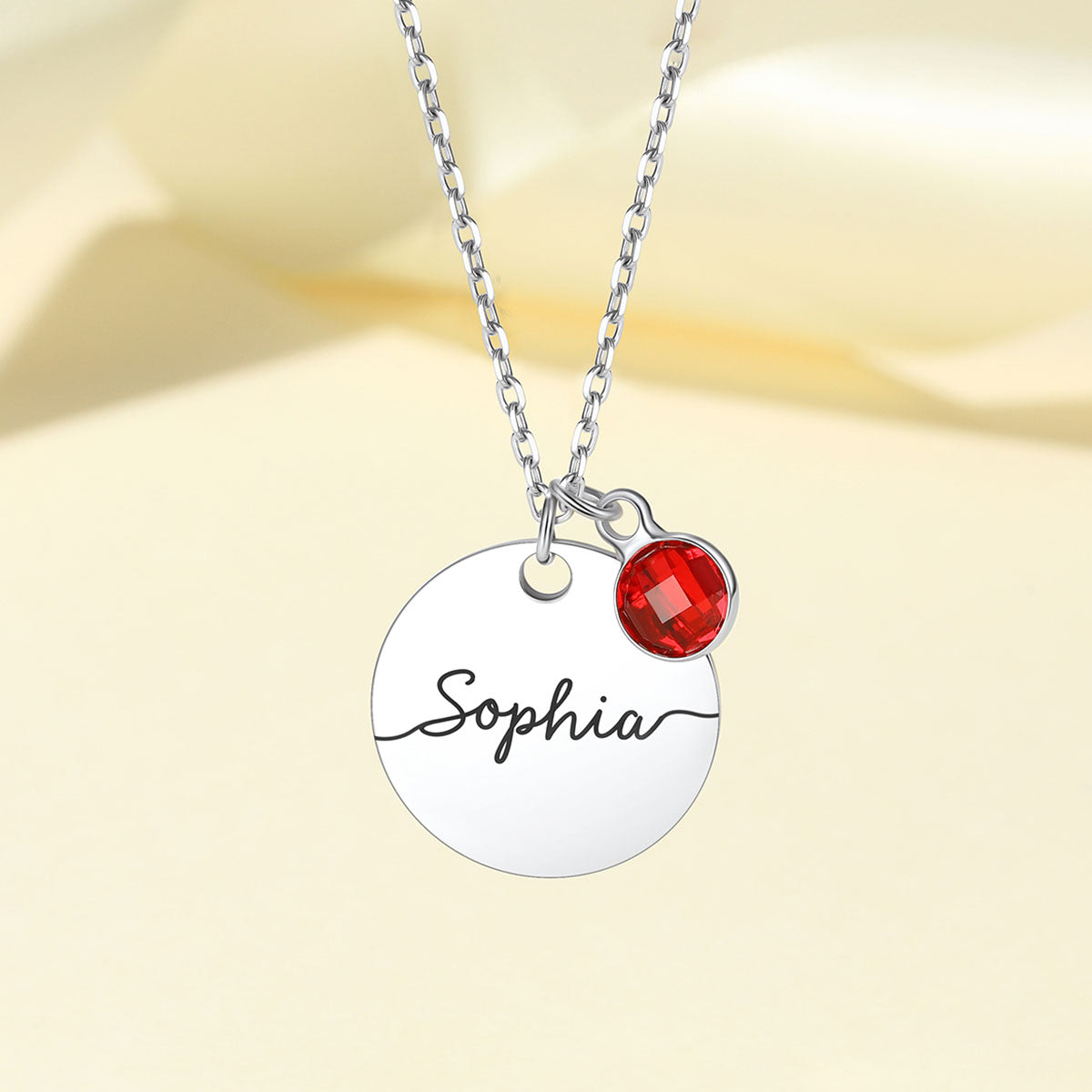 A silver-tone necklace with a round engraved disc pendant reading 'Sophia' and a small faceted red crystal charm, shown on a soft cream background.