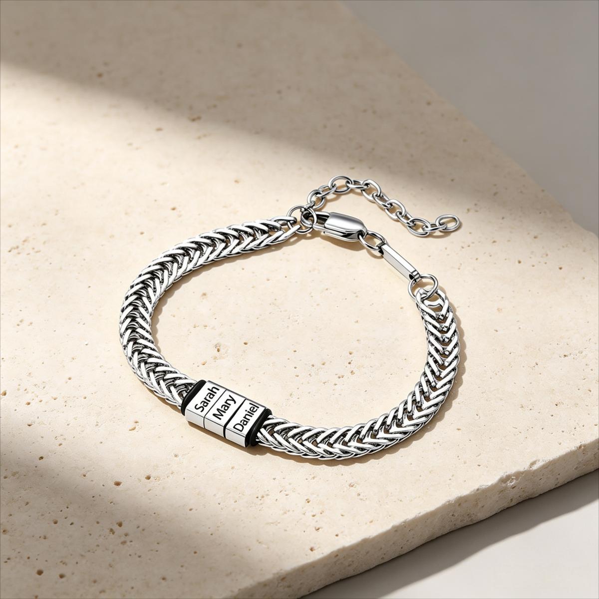 A silver-toned braided chain bracelet with a rectangular engraved plate reading 'Sola Deo', displayed on a light beige stone surface with soft shadows.