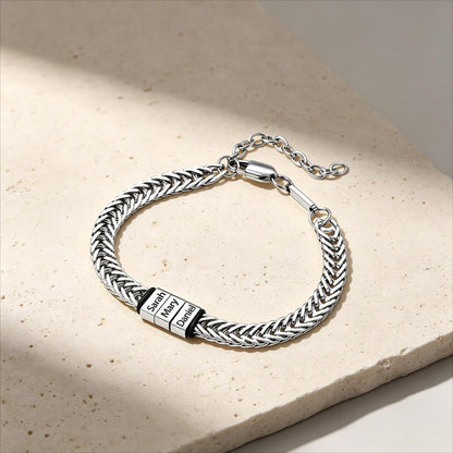 A silver-toned braided chain bracelet with a rectangular engraved plate reading 'Sola Deo', displayed on a light beige stone surface with soft shadows.