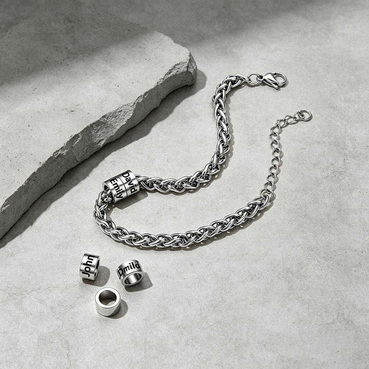 A silver-toned chain bracelet with three cylindrical bead charms engraved with 'HIT' is arranged on a light gray textured surface next to a rough stone slab. The clasp is open, and the three charms are placed separately below the bracelet.