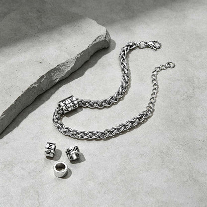 A silver-toned chain bracelet with three cylindrical bead charms engraved with 'HIT' is arranged on a light gray textured surface next to a rough stone slab. The clasp is open, and the three charms are placed separately below the bracelet.