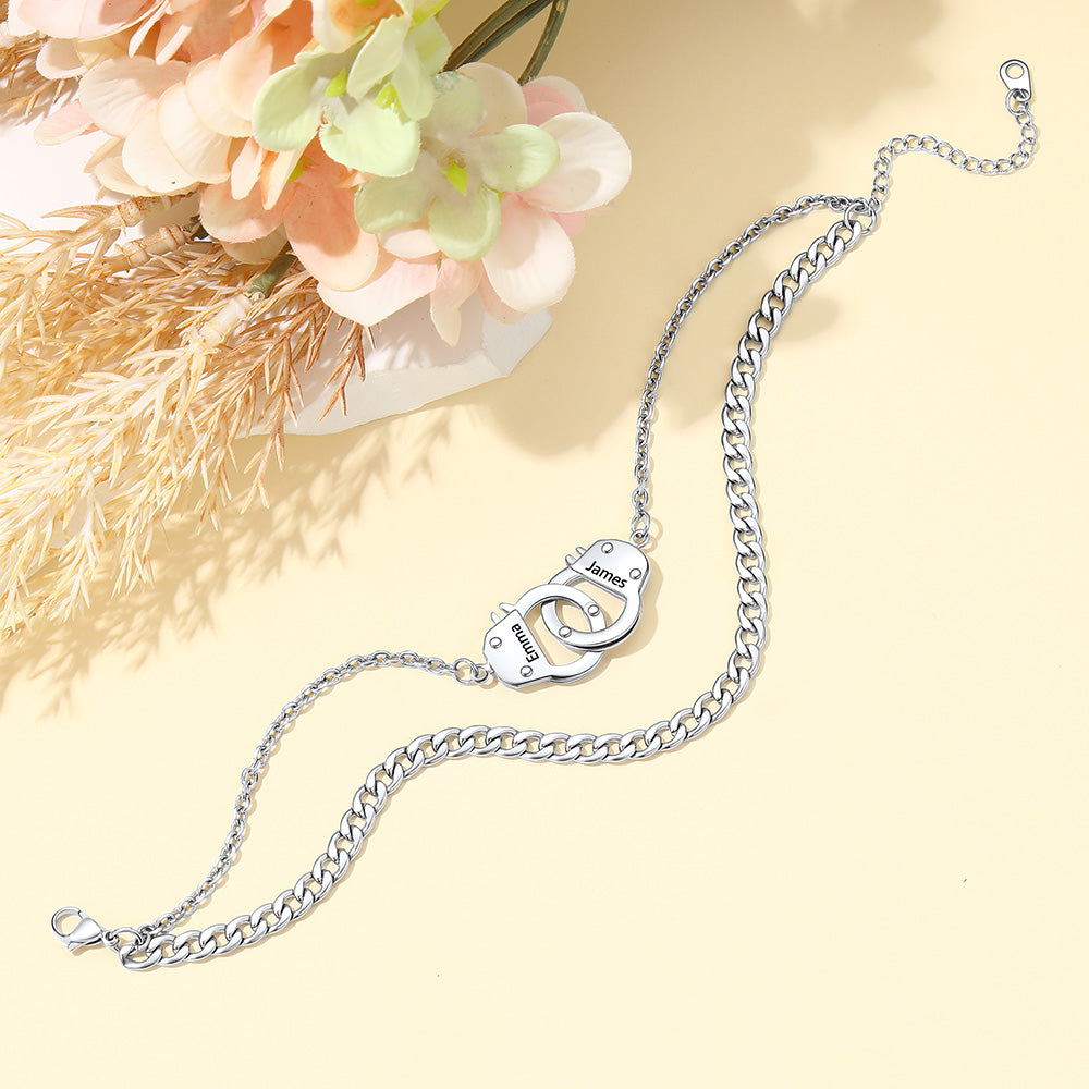 A silver-toned chain handcuff anklett with a unique interlocking clasp shaped like two hands, displayed on a light beige background with decorative flowers in the corner.