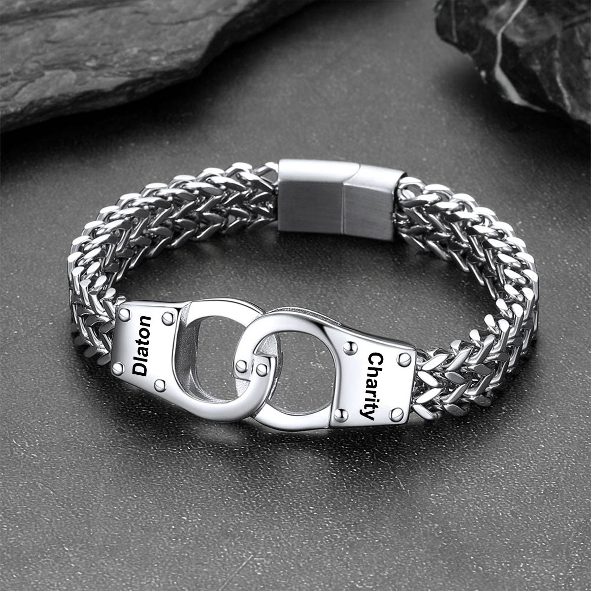 A silver-toned stainless steel chain bracelet with a unique clasp design resembling two interlocking handcuffs. The clasp features engraved text reading 'Diaton' on one cuff and 'Charity' on the other, set against a dark textured background with slate props.