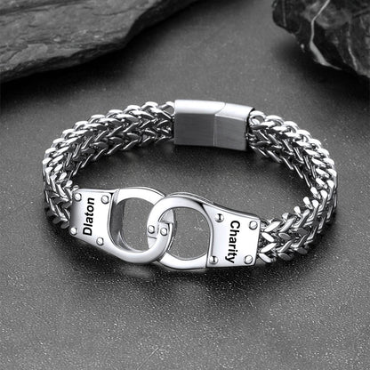 A silver-toned stainless steel chain bracelet with a unique clasp design resembling two interlocking handcuffs. The clasp features engraved text reading 'Diaton' on one cuff and 'Charity' on the other, set against a dark textured background with slate props.