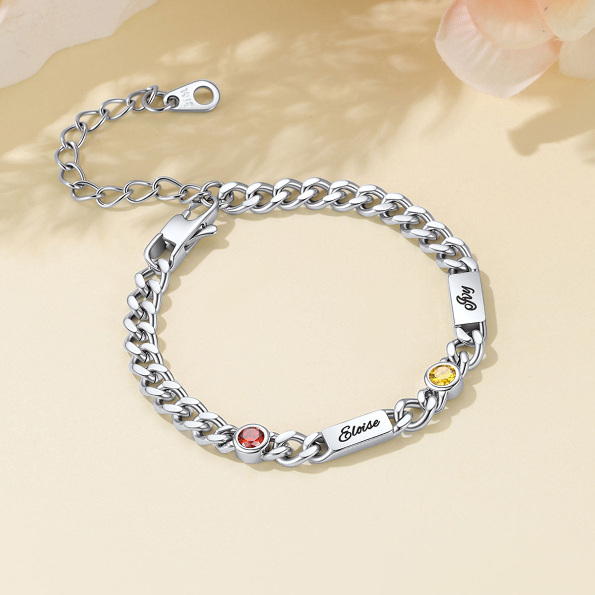 A silver Cuban chain bracelet with three engraved name plates, each set with a small round gemstone in red, yellow, and pink, displayed on a warm beige background.