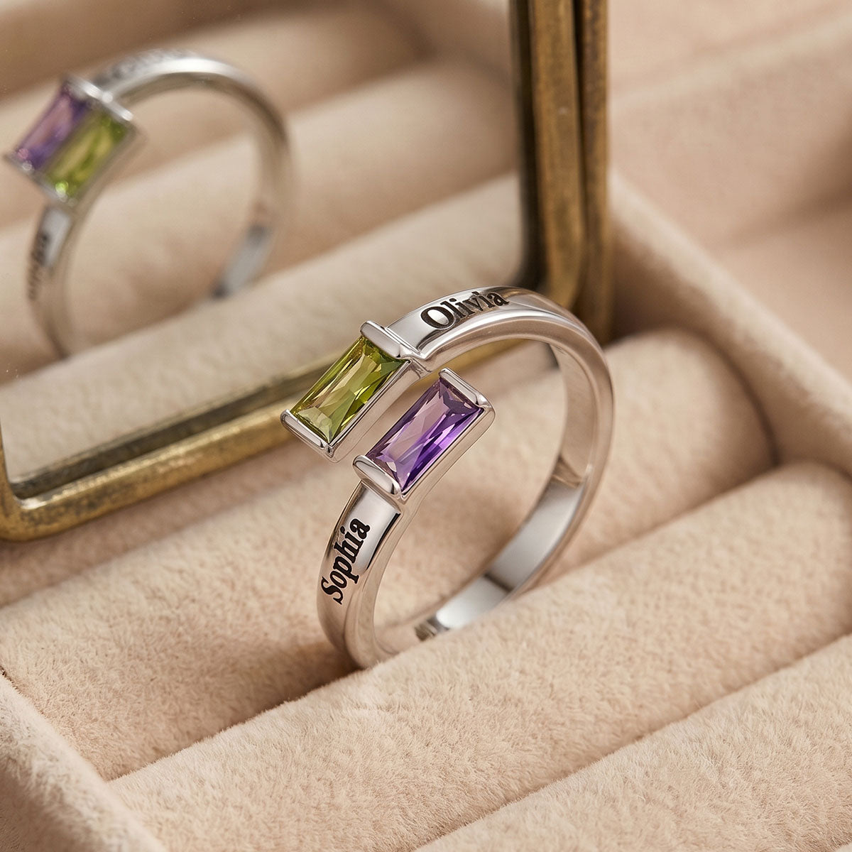 A silver baguette birthtone ring with a split design featuring a green topaz and a purple amethyst rectangular gemstone, personalized with the names 'Olivea' and 'Sophia' engraved on the band. The ring is displayed in a beige jewelry box with a mirror reflecting its back.