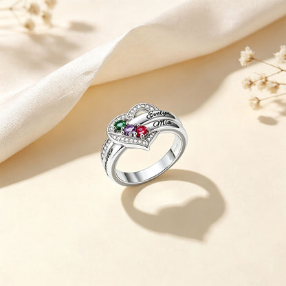 A silver birthstone heart ring with colorful gemstones and the engraved names 'Evelyn' and 'Mia', styled on a beige surface with a cream fabric drape and small dried flowers.