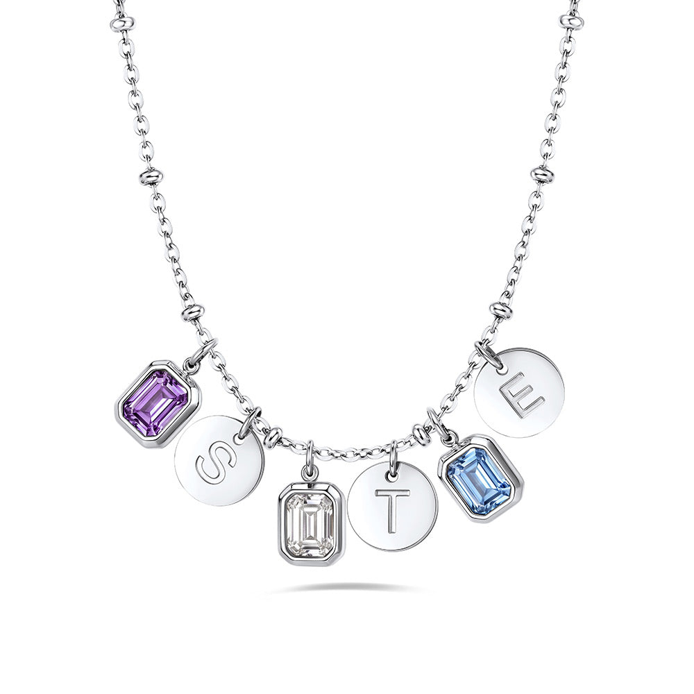A silver birthstone initial necklace featuring five dangling charms: two rectangular purple and light blue emerald-cut gemstones, two round silver discs engraved with the letters S and T, and one round silver disc engraved with the letter E. The necklace is displayed on a clean white background.