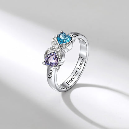 A silver birthstone ring with a twisted design featuring a light blue heart-shaped gemstone and a purple pear-shaped gemstone, accented with small clear stones. The inner band is engraved with 'Mary' and 'Forever Love'.