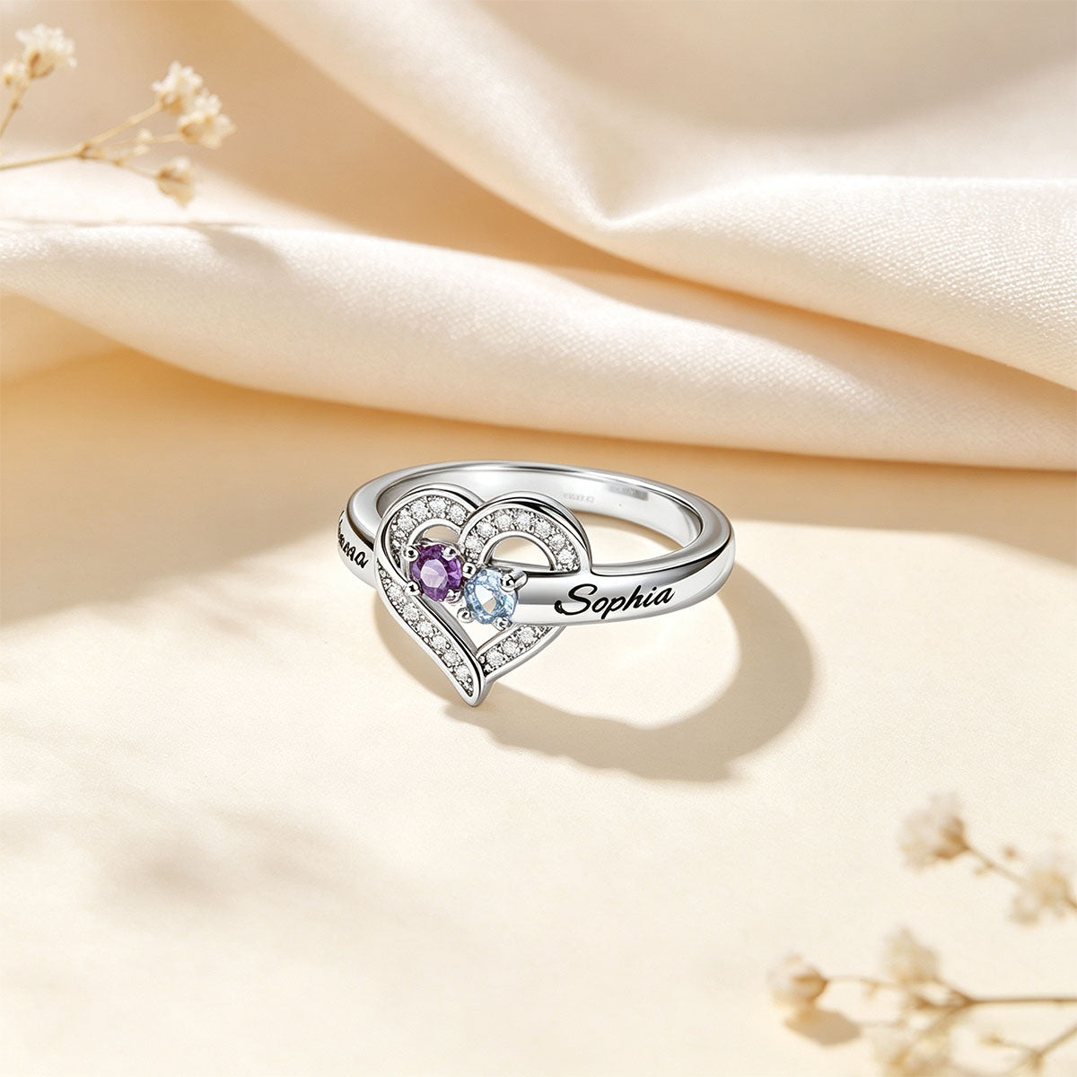 A silver birthstone ring with clear stones and a purple center stone, featuring the name 'Sophia' engraved on one side, displayed on a beige satin fabric background with dried flowers.