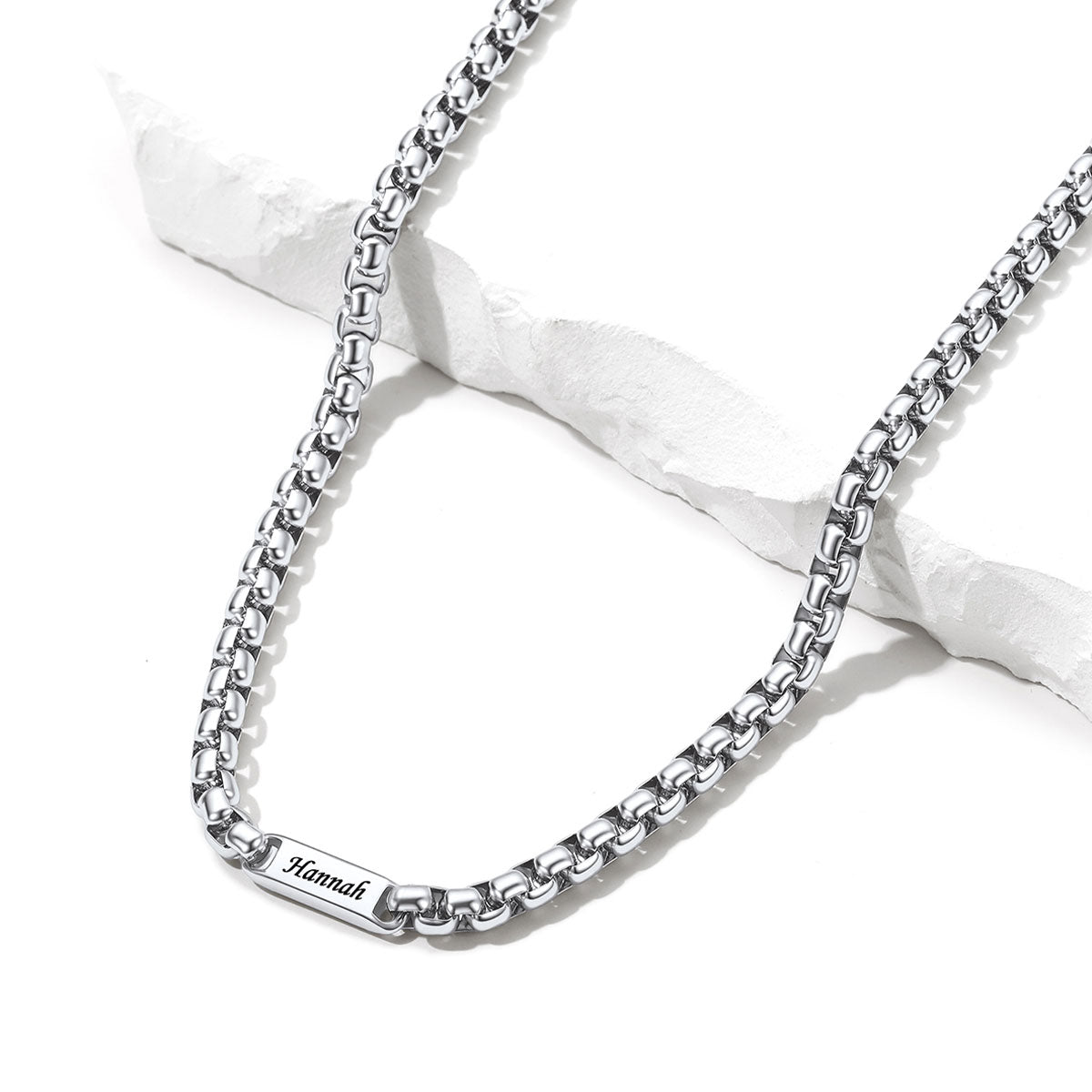 A silver box chain name necklace with a rectangular engraved plate reading 'Mannah', displayed on a white textured stone prop against a white background.