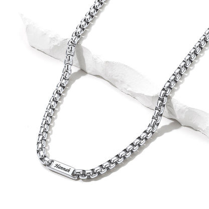 A silver box chain name necklace with a rectangular engraved plate reading 'Mannah', displayed on a white textured stone prop against a white background.