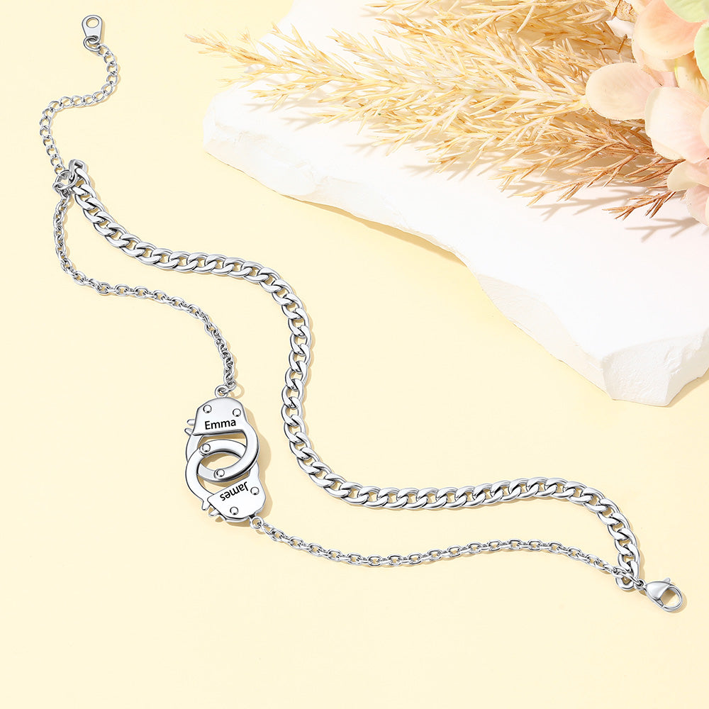 A silver chain ankle bracelet with a custom-engraved padlock pendant that reads 'Emma' and 'Love', styled on a pale yellow background with dried grass and a white stone prop.