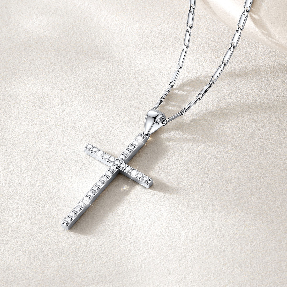 A silver cross necklace adorned with small clear stones, resting on a textured beige surface with a soft cream fabric corner in the background.