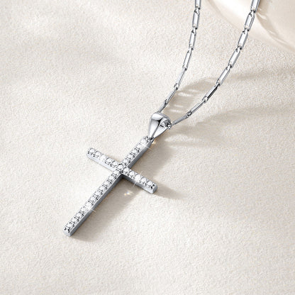 A silver cross necklace adorned with small clear stones, resting on a textured beige surface with a soft cream fabric corner in the background.