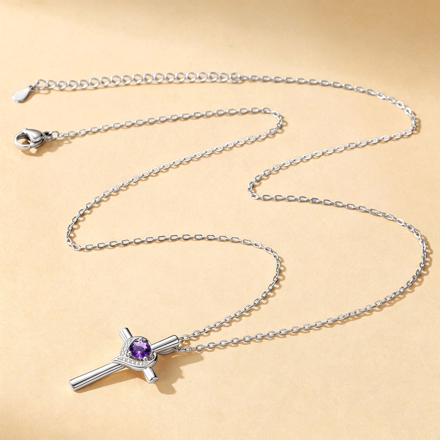 A silver cross pendant necklace featuring a central purple gemstone surrounded by a halo of small clear stones, displayed on a warm beige background.