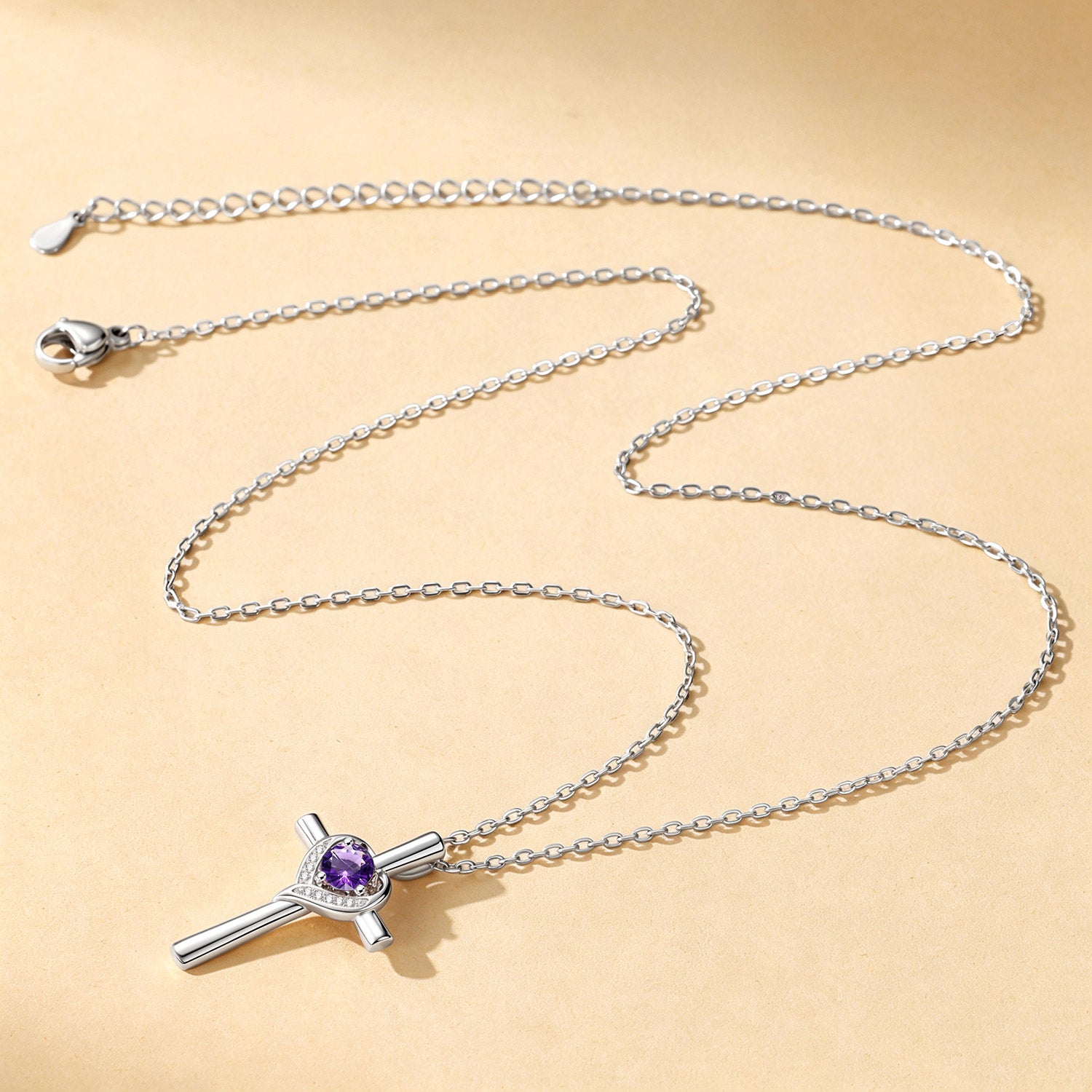 A silver cross pendant necklace featuring a central purple gemstone surrounded by a halo of small clear stones, displayed on a warm beige background.
