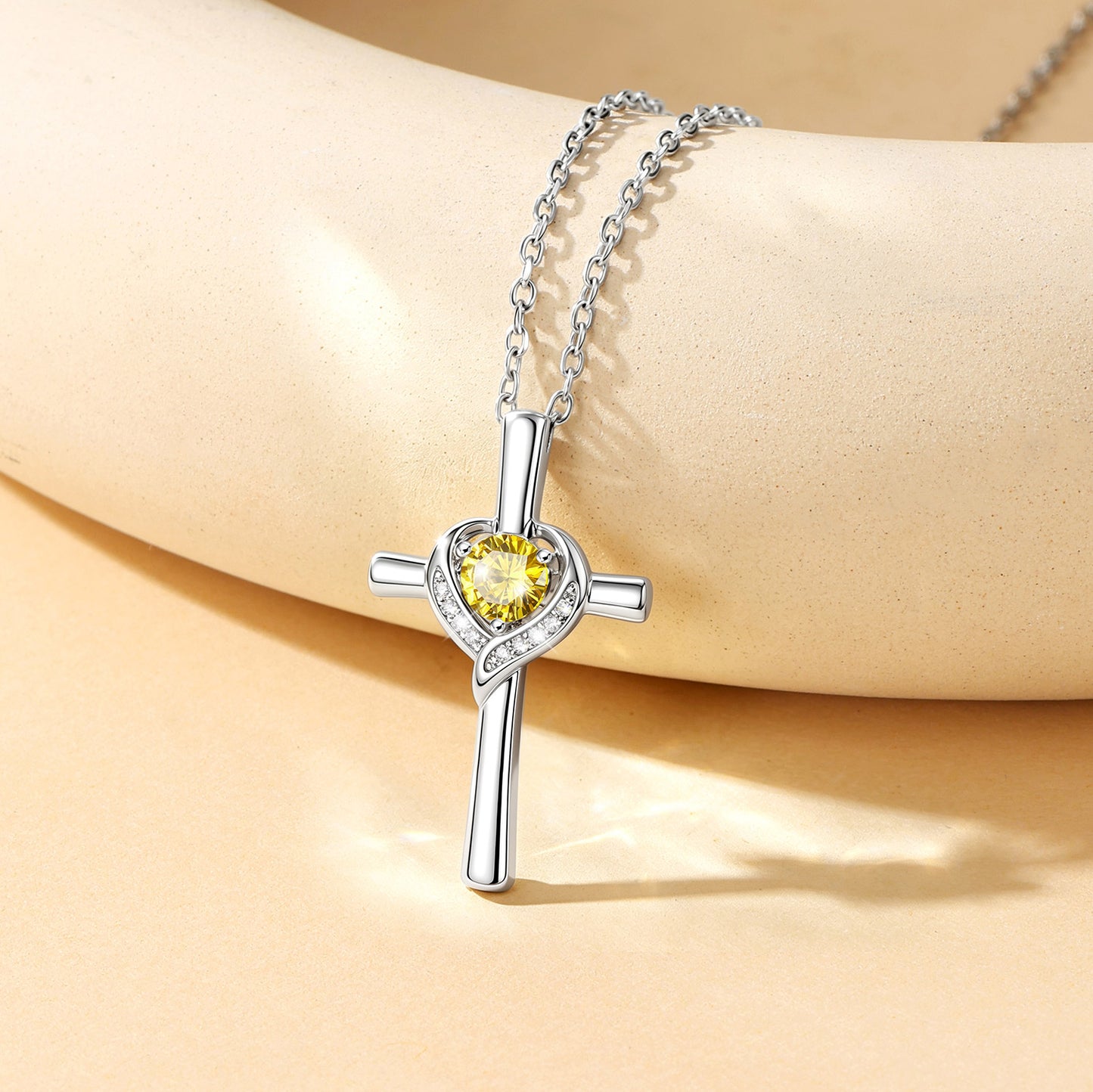 A silver cross pendant necklace with a yellow heart-shaped crystal and small clear stones, displayed on a beige mannequin arm.