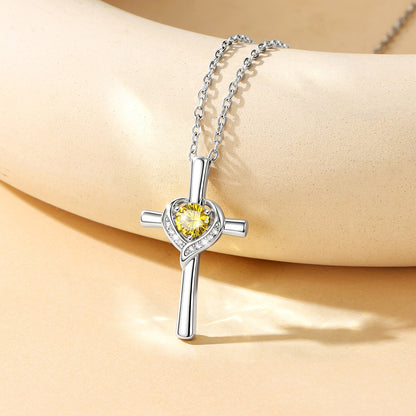 A silver cross pendant necklace with a yellow heart-shaped crystal and small clear stones, displayed on a beige mannequin arm.