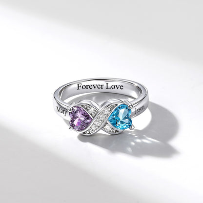 A silver infinity birthstone ring featuring a heart-shaped light blue gemstone and a purple heart-shaped gemstone, accented with small clear stones. The band is engraved with 'Forever Love' and names 'Mary' and 'Jason'.