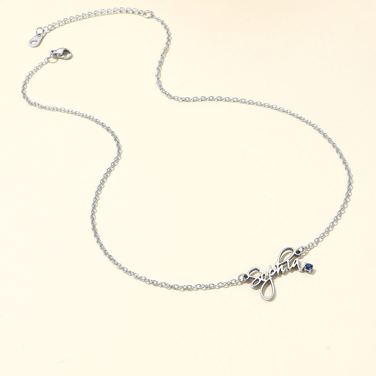 A silver name necklace featuring a cursive script nameplate pendant and a small blue stone accent, displayed on a light cream background.