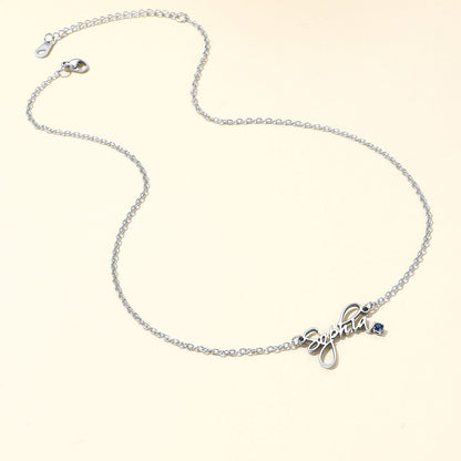 A silver name necklace featuring a cursive script nameplate pendant and a small blue stone accent, displayed on a light cream background.