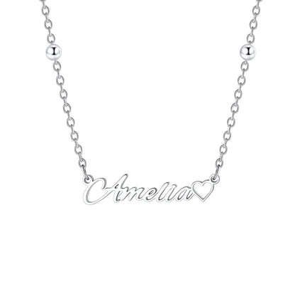 A silver name necklace featuring a script nameplate that reads 'Amelia' in cursive, with a small heart outline at the end of the word, shown on a plain white background.