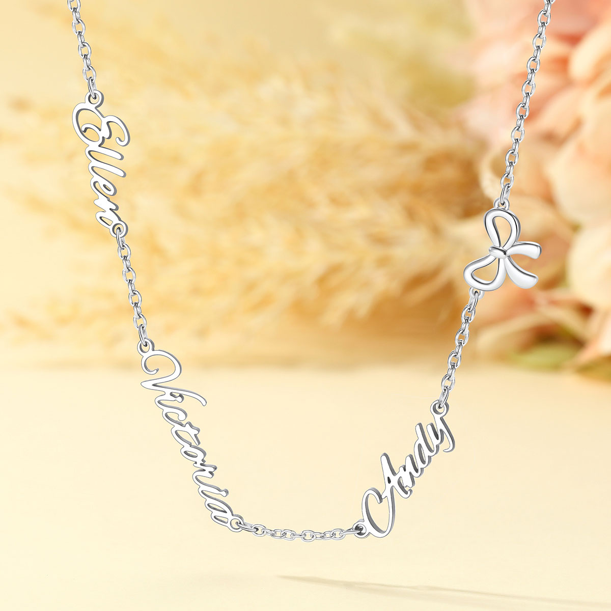 A silver name necklace featuring the script names 'Eve', 'Admirer', and 'Andy' connected by a delicate chain, with a small bow charm on the right side, shown against a soft beige background with blurred flowers.