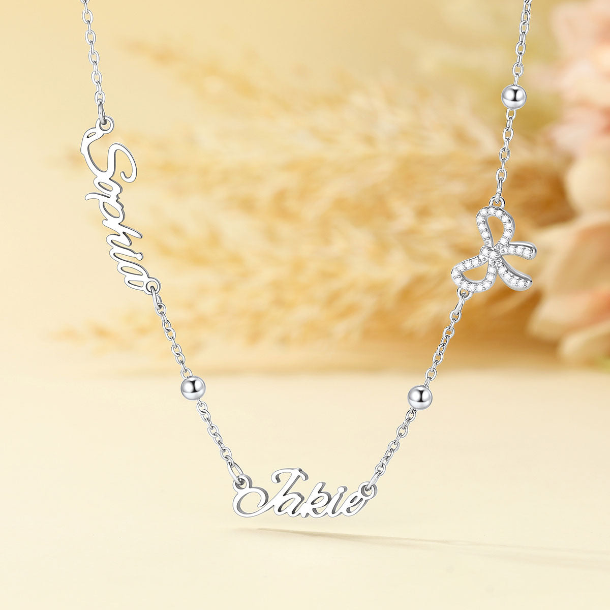 A silver name necklace with custom script lettering, featuring a nameplate at the bottom and a small bow charm embellished with clear rhinestones, displayed on a soft beige background.