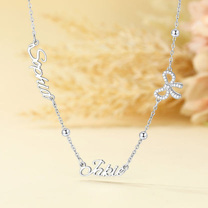 A silver name necklace with custom script lettering, featuring a nameplate at the bottom and a small bow charm embellished with clear rhinestones, displayed on a soft beige background.